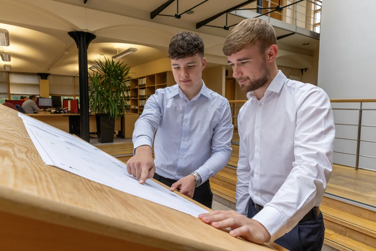 Two people leaning over a large drawing spread out on a table in an office, one of them pointing at a detail on the sheet.