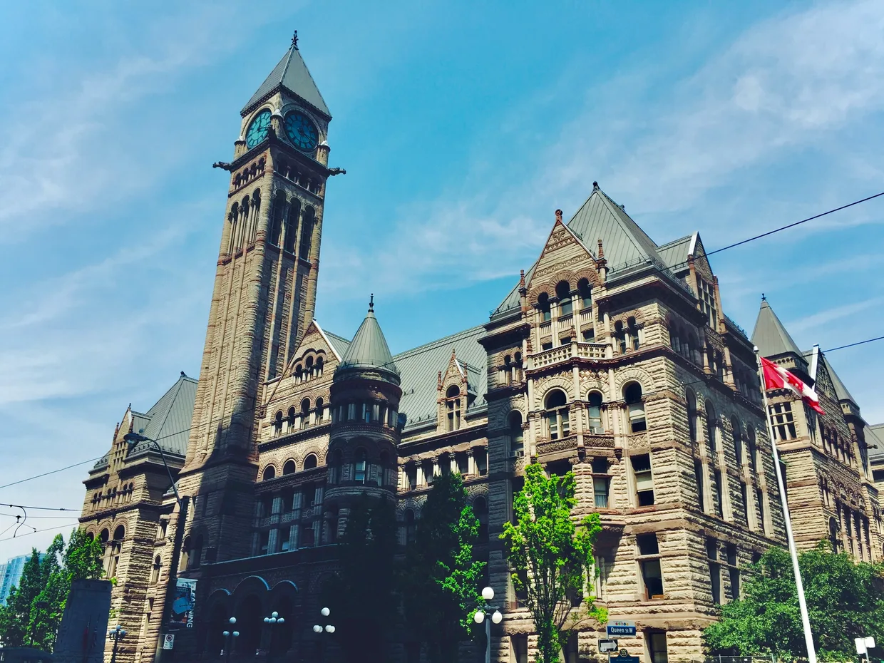 An old city hall from the street below, its clock tower and stone facade against a clear sky with a Canadian flag flying beside it.