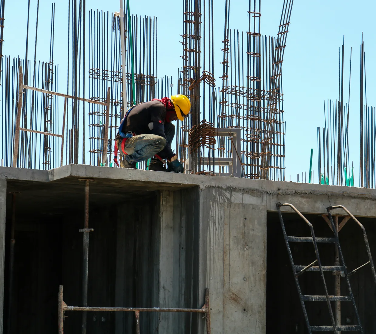 A worker in a hard hat crouched on the edge of a concrete deck, tying reinforcing steel that stands up in bundles behind them.