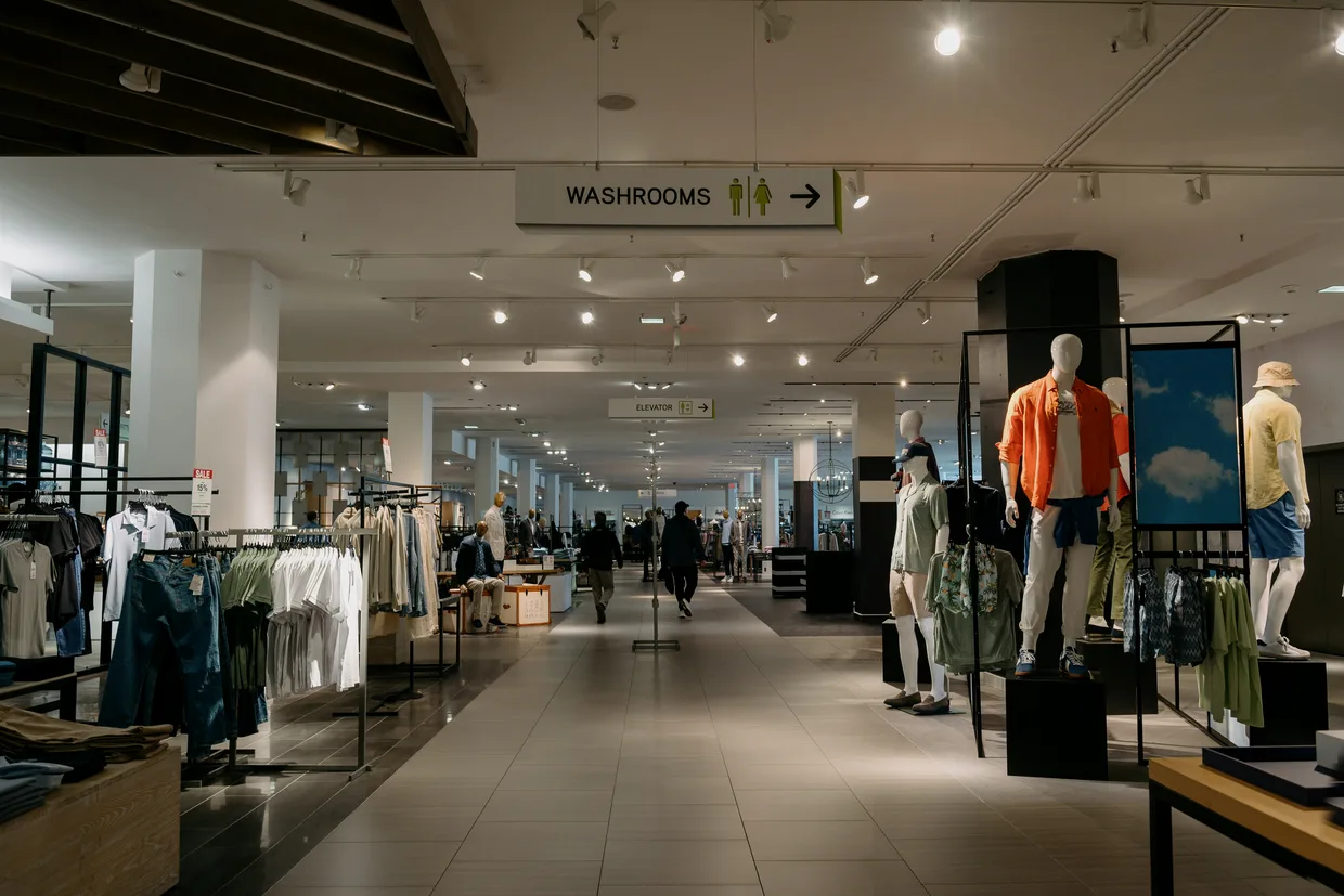 A finished retail floor, a wide aisle running away between clothing racks and mannequins with an elevator sign hung over it.