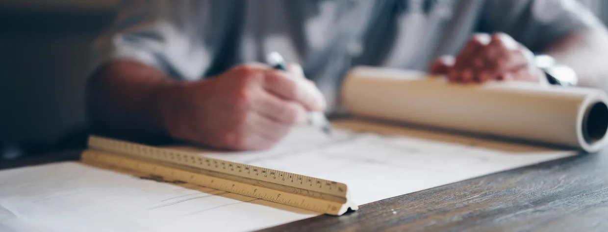 Hands drawing on a sheet at a desk with a pen, a wooden scale rule laid across the drawing and a rolled up set beside it.